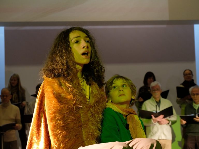 Sängerin im Kostüm mit dem kleinen Prinzen, im Hintergrund der Chor